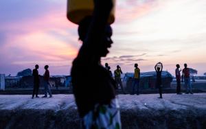  woman walks past a group of young men after collecting water just as the sun set inside the UN Protection of Civilians site in Bentiu. [ © Dominic Nahr / MAPS] 