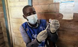 These are vaccinators at the Kihindo vaccination site in the Kirotshe Health Zone. In front of them are gloves and syringes. One of the vaccinator removes the vaccine from the cold chain and draws it into a syringe. They then hand it to a colleague who administers the vaccination. 