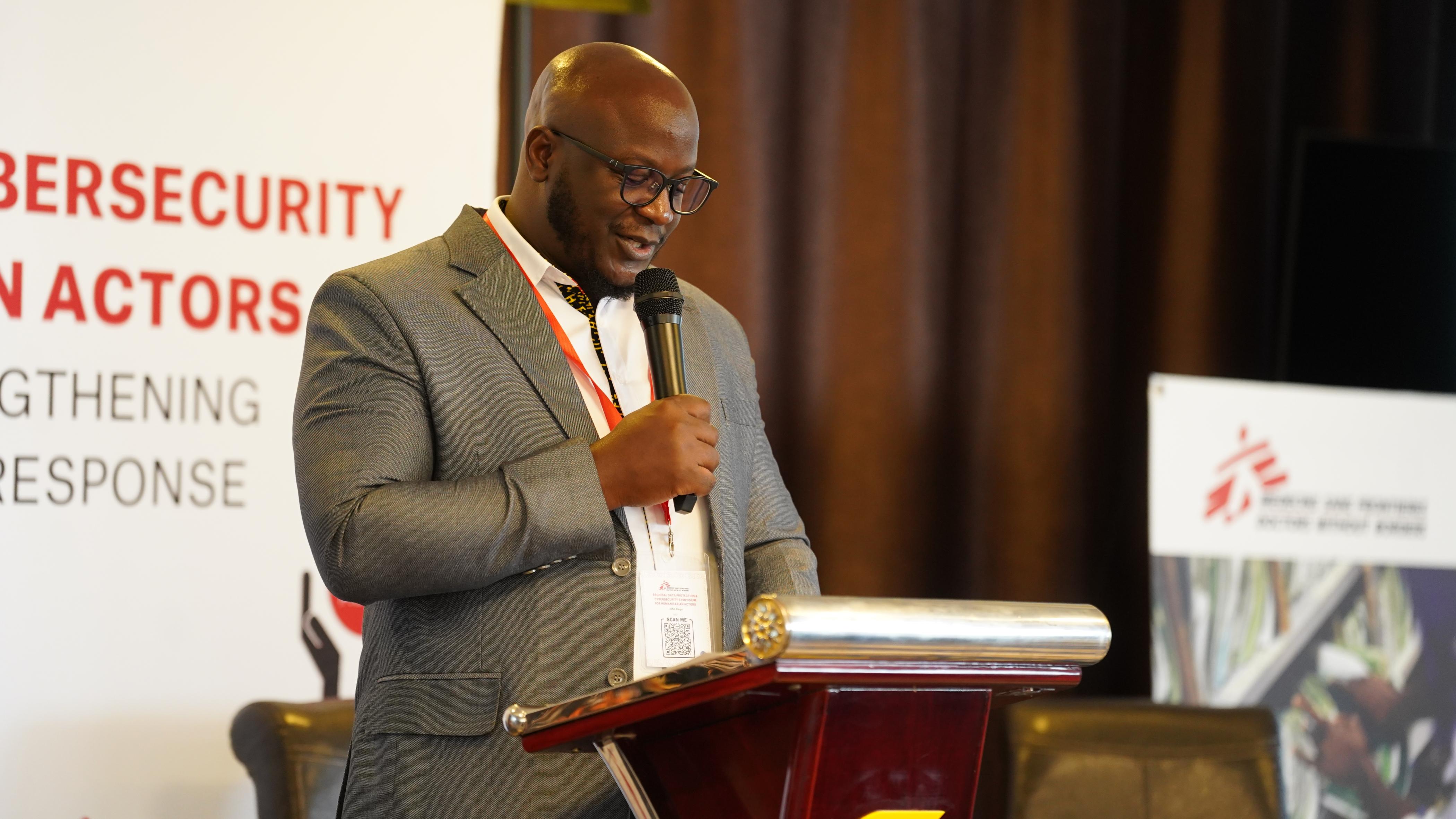 John Riaga standing at a podium addressing attendees at the Regional Data Protection and Cybersecurity Symposium for Humanitarian Actors.