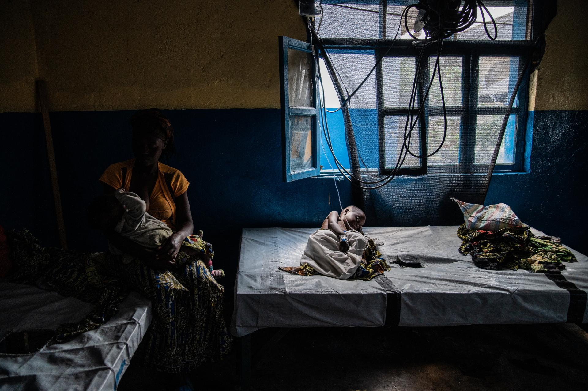 A woman sits with her child in her arms as her youngest son lies in bed while being treated for cholera at the Sake Referral Center, supported by MSF as part of its emergency response. They had to leave the Bulengo displacement camp to come to Sake