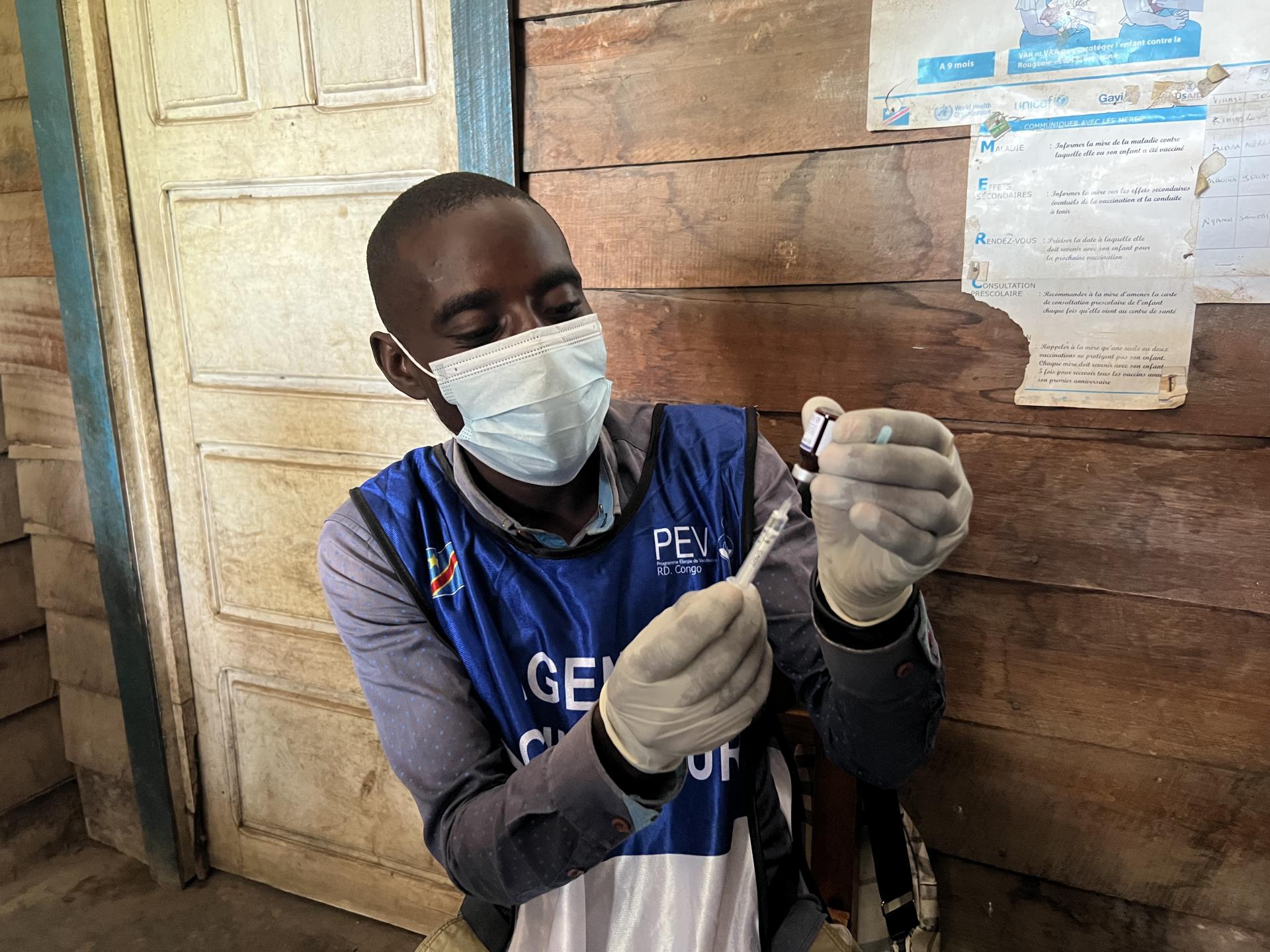 These are vaccinators at the Kihindo vaccination site in the Kirotshe Health Zone. In front of them are gloves and syringes. One of the vaccinator removes the vaccine from the cold chain and draws it into a syringe. They then hand it to a colleague who administers the vaccination. 