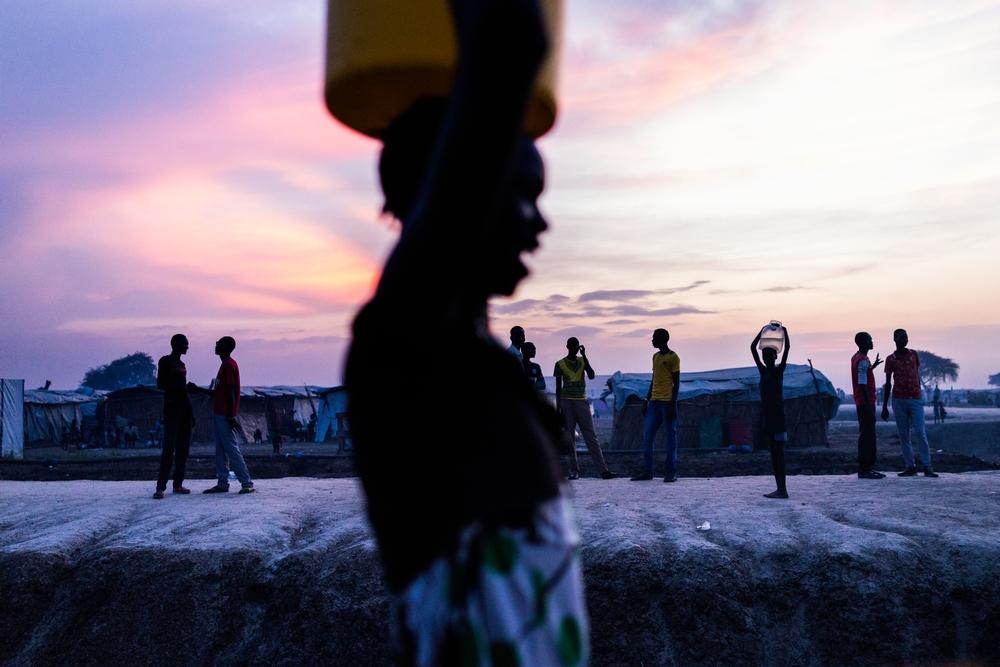  woman walks past a group of young men after collecting water just as the sun set inside the UN Protection of Civilians site in Bentiu. [ © Dominic Nahr / MAPS] 