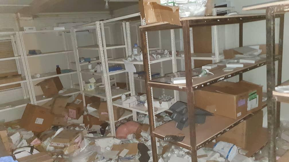 A wide shot of a severely ransacked room, likely a storage or pharmacy area, with shelves completely emptied or holding only scattered debris. The floor is littered with torn packaging, broken items, and cardboard boxes, conveying a scene of catastrophic loss due to looting.