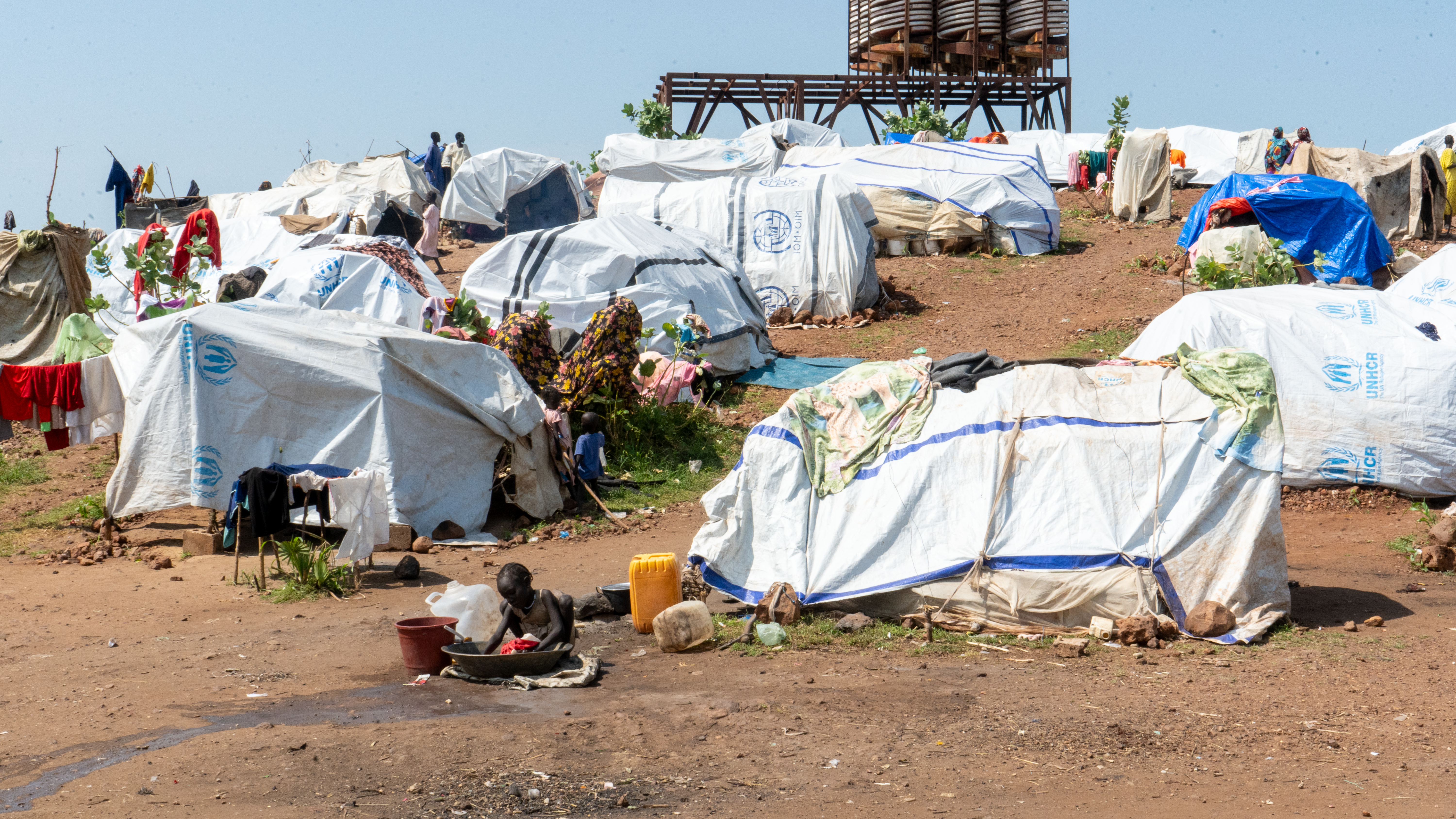 A young girl bends over a basin, washing clothes outside her family’s small shelter.